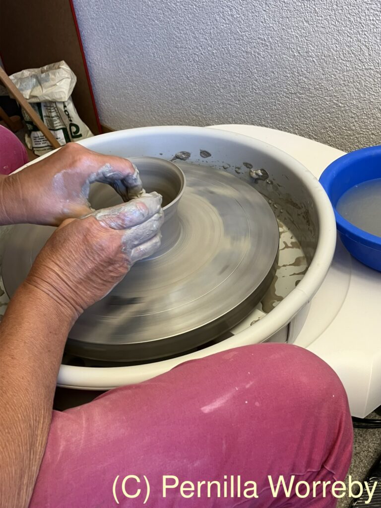 Eine Person formt ein kleines Keramikwarenstück auf einer Drehscheibe, während ihre Hände mit Ton und Wasser bearbeitet werden.